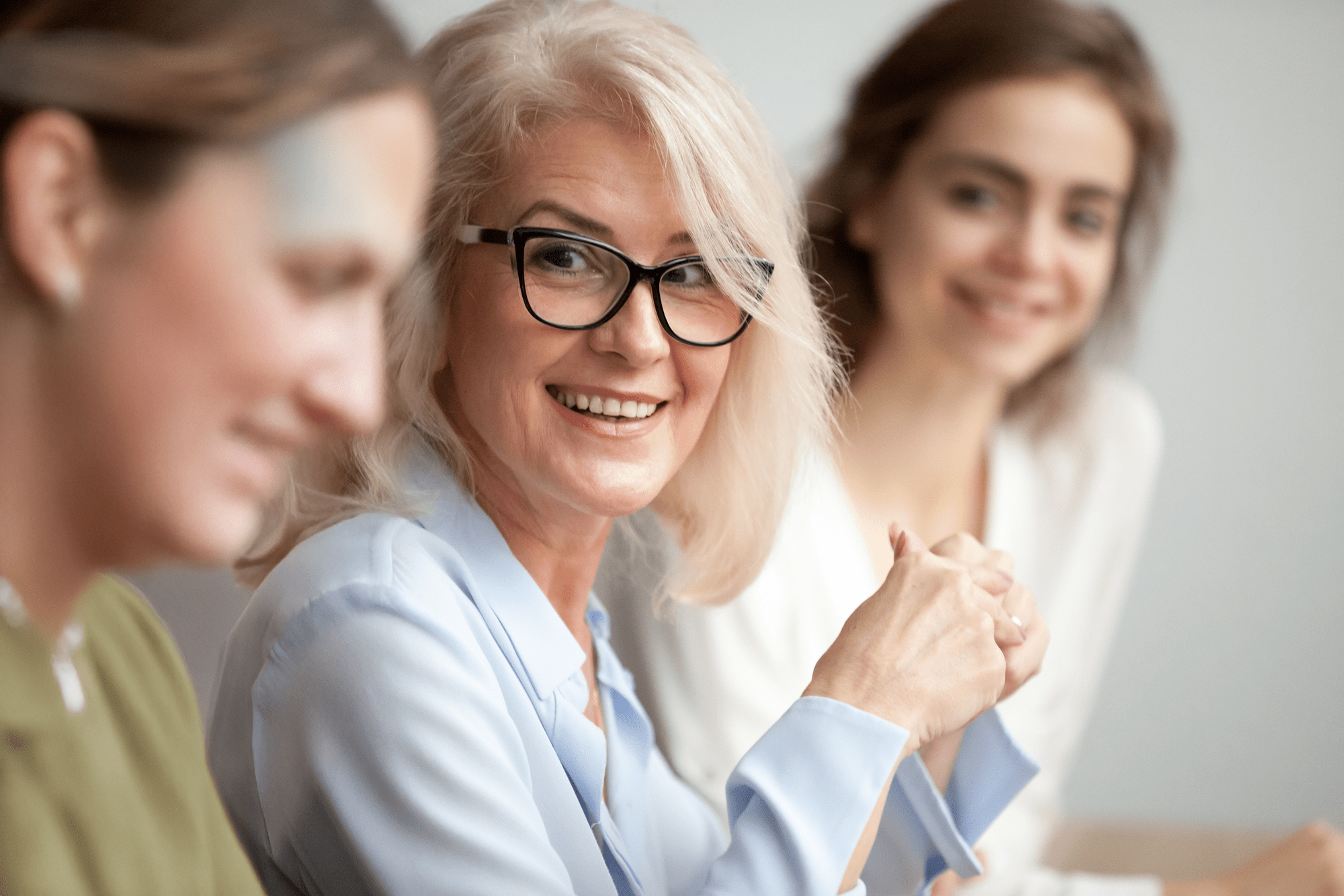 Three women in a meeting, with a focus on a smiling older woman in a light blue blouse and glasses, engaged in conversation.