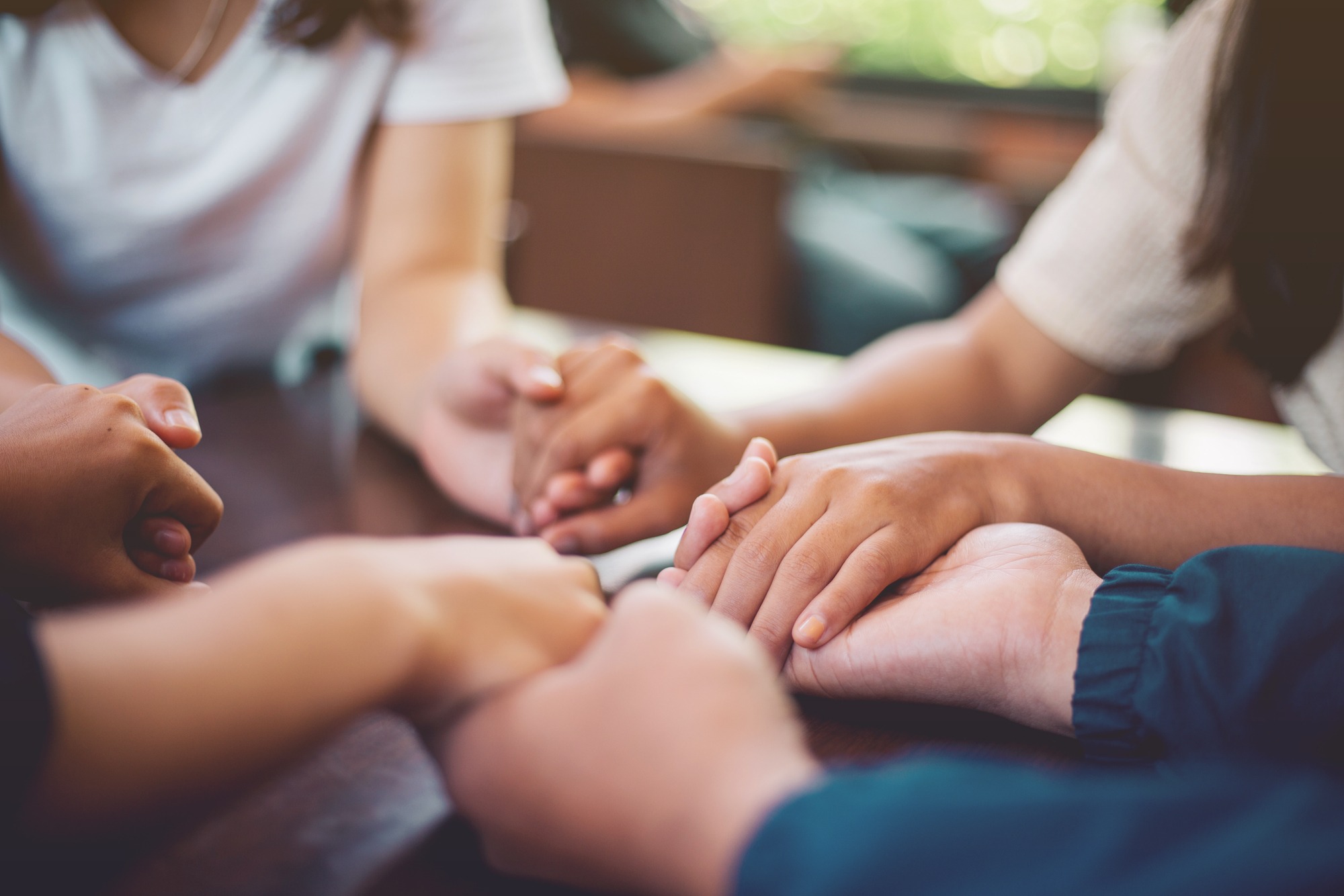 Multiple hands clasp together on a table, symbolizing unity and support in a group setting, likely during a discussion or meeting.