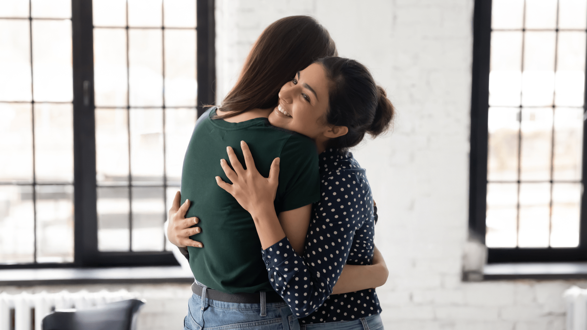 Two women embrace warmly in a cozy, sunlit room, showcasing friendship and support in a moment of joy and connection.