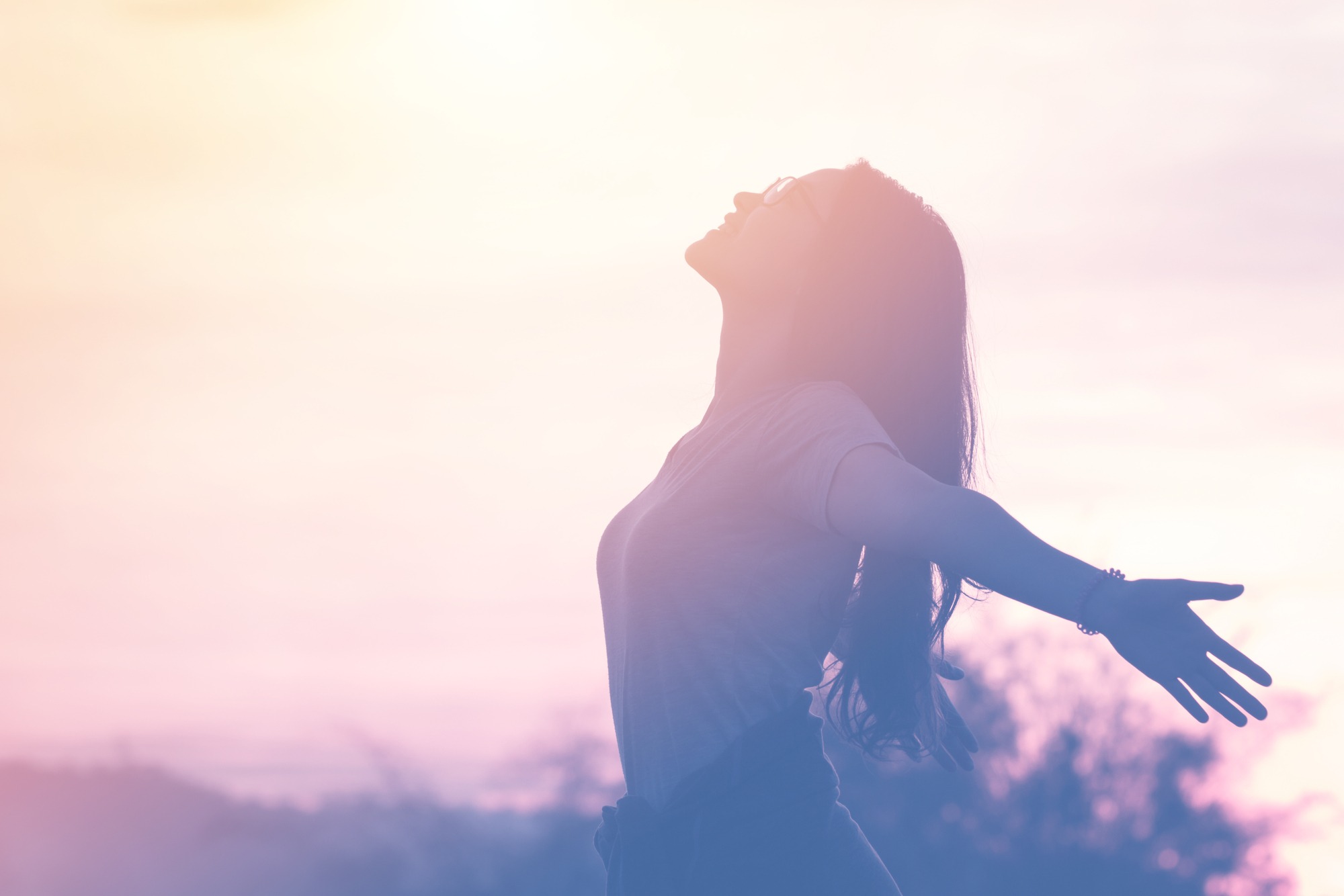 A woman with long hair embraces the sunset, arms outstretched, symbolizing freedom and positivity against a soft, colorful sky.