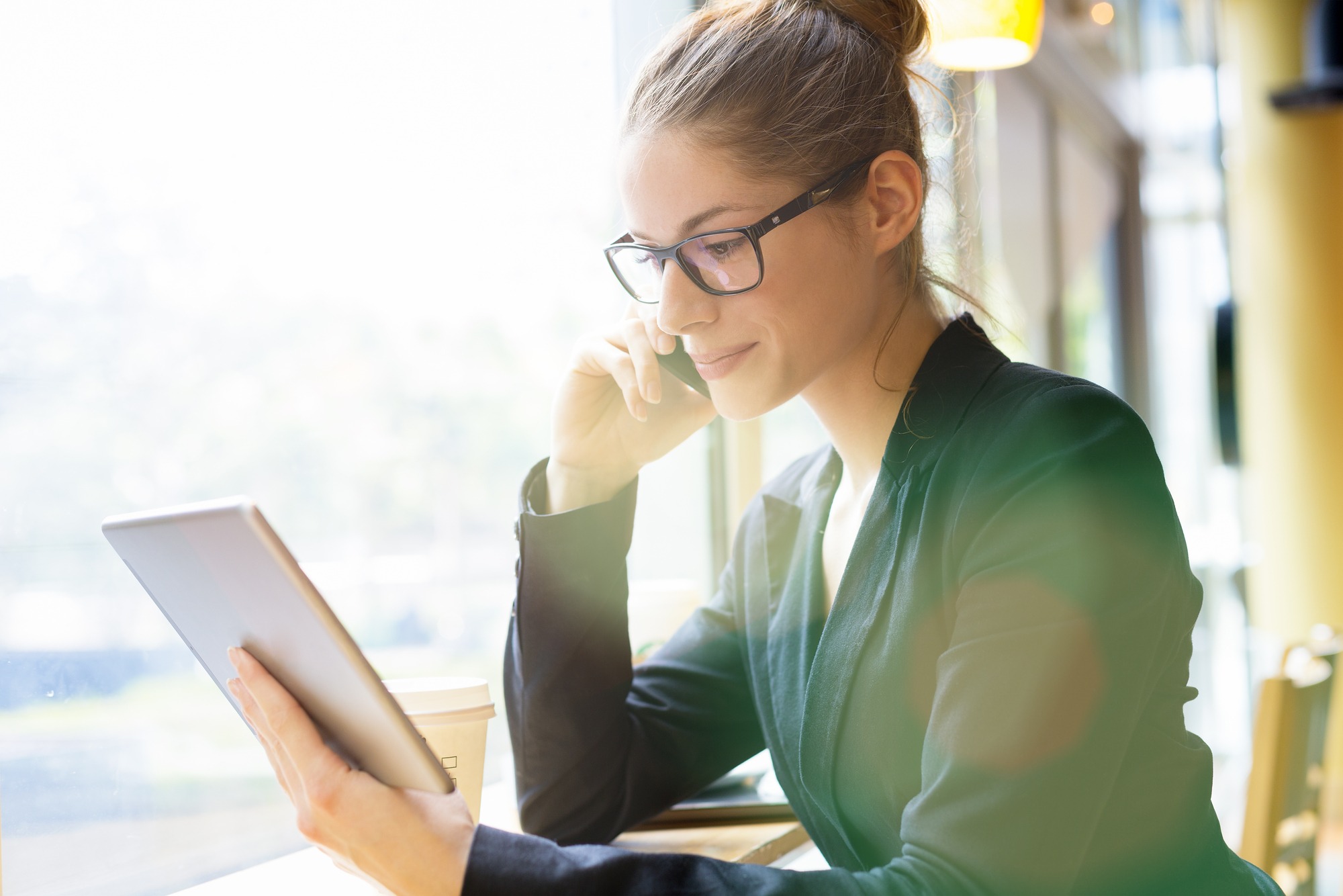Professional woman in a blazer uses a tablet while talking on the phone, seated at a café with a coffee cup nearby.